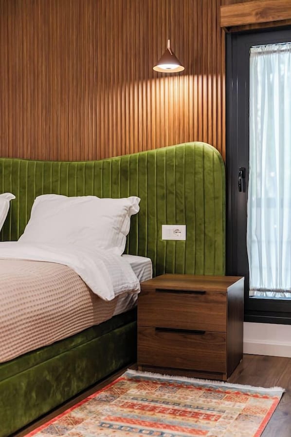 Un dormitorio con cabecera tapizada en verde, una mesita de noche de madera y una alfombra estampada.