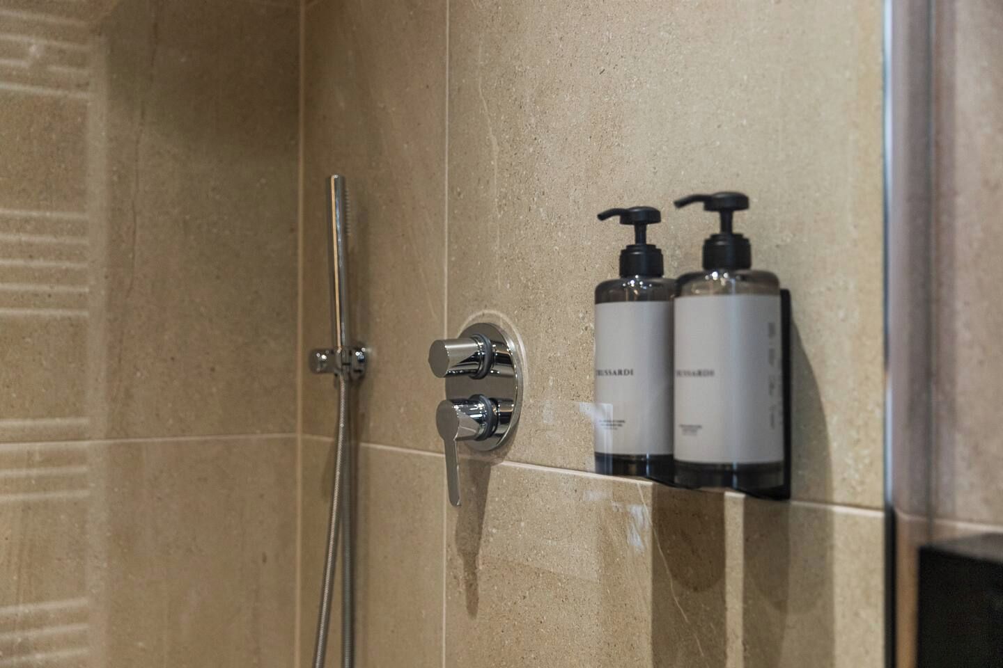 A shower with a chrome faucet and a handheld showerhead. There are two bottles of body lotion on the wall.