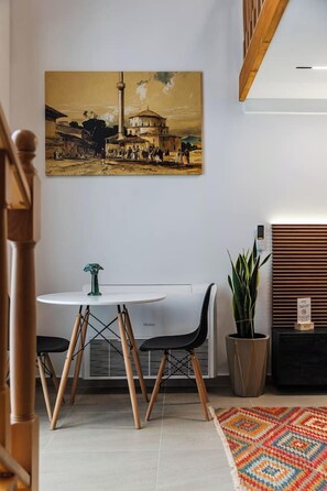 A room with a white table, black chairs, a potted plant, a colorful rug, and a painting on the wall. - Muraga Villas 6 | Sofra (Tiranë)