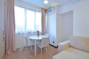 Ein modernes Wohnzimmer mit einem runden Esstisch, zwei Stühlen, einer Couch und einem Fenster mit leichtem Vorhang.