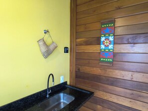 A kitchen with a black countertop, a stainless steel sink, a woven basket hanging on the wall, and a colorful decorative piece. - Alma Zen House - Comfort and beach (180m) Air Conditioning/Heated Pool (Guaratuba)