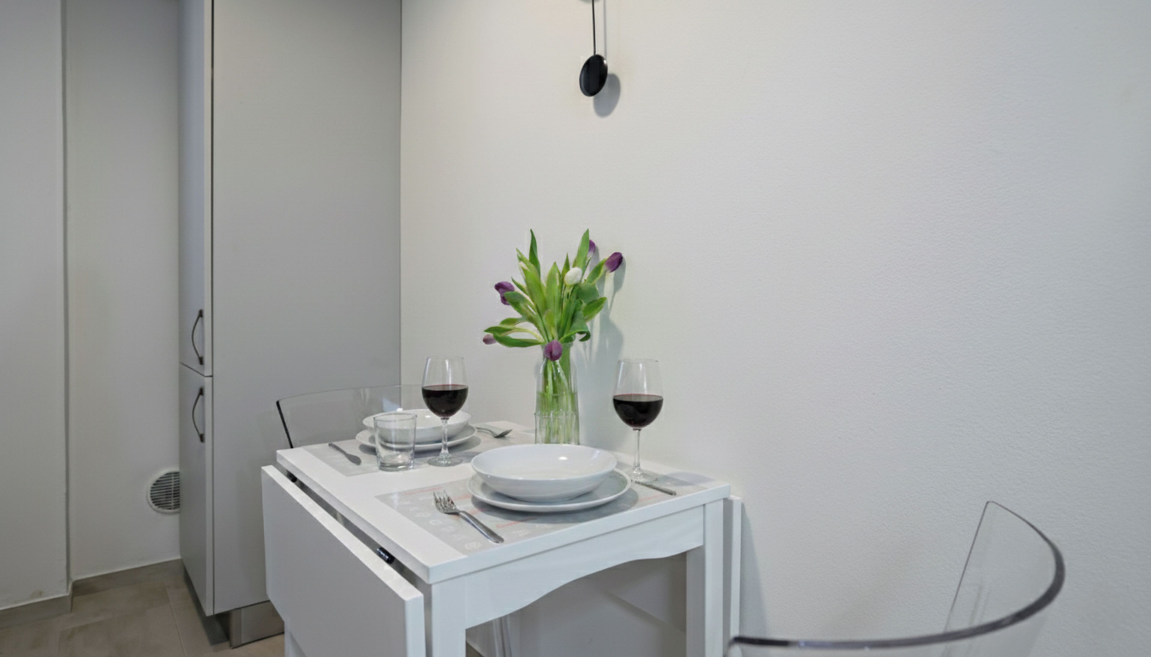 Ein kleiner, moderner Essbereich mit einem weißen Tisch, gedeckt für zwei Personen, mit einer Blumenvase und zwei Gläsern Rotwein.