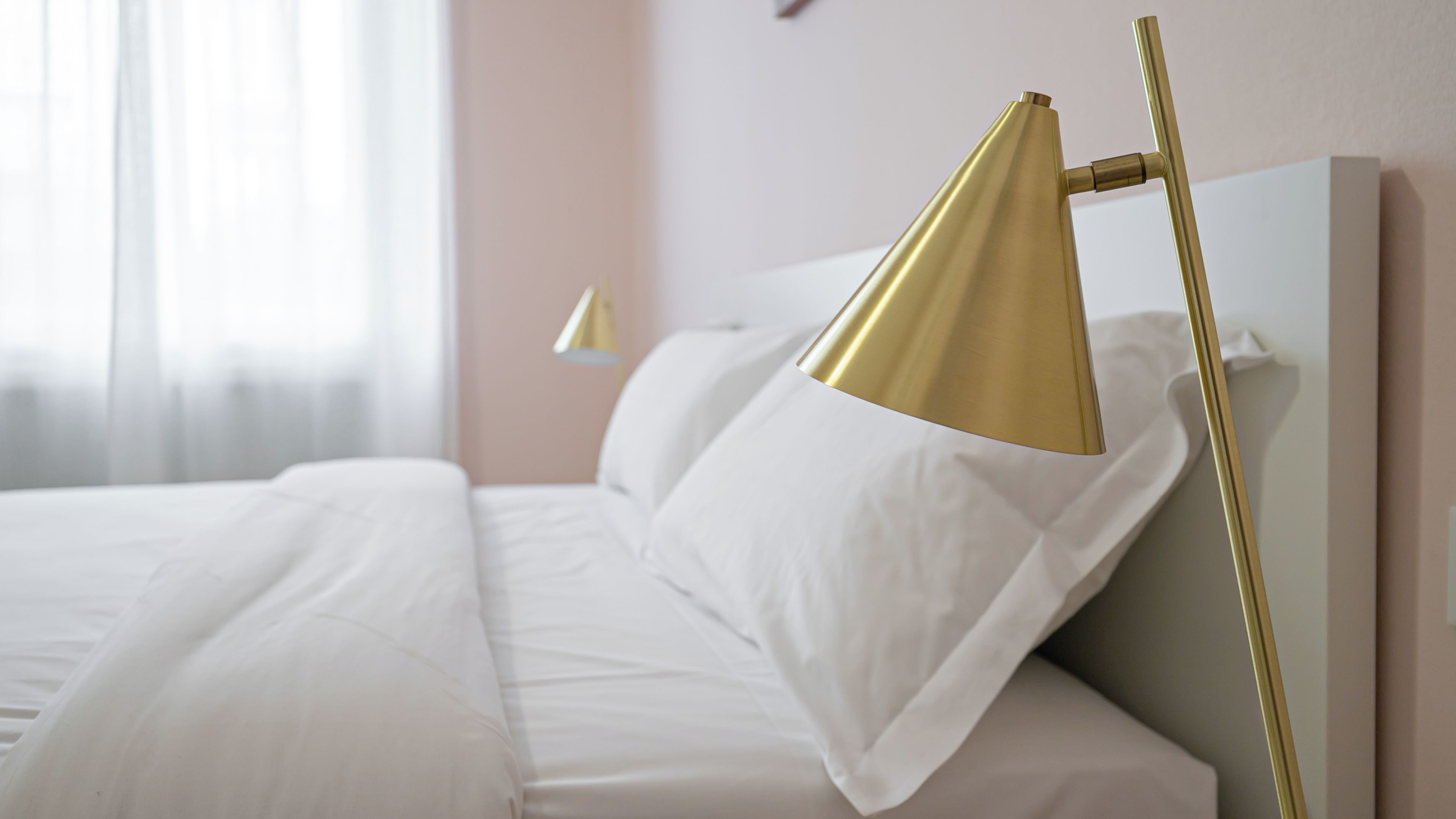 Ein ordentlich bezogenes Bett mit weißen Leinen, eine goldfarbene Lampe daneben und eine weiße Wand im Hintergrund.