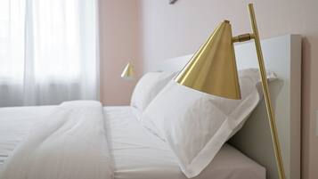 Ein ordentlich bezogenes Bett mit weißen Leinen, eine goldfarbene Lampe daneben und eine weiße Wand im Hintergrund.
