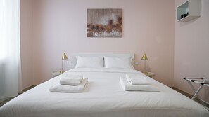 Een netjes opgemaakt bed met witte lakens en twee handdoeken gevouwen aan het voeteneinde. Twee bedlampen staan op witte nachtkastjes. Daarboven hangt een schilderij.