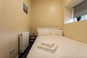 Una habitació d'hotel amb un llit, un radiador, una taula petita amb una planta i un quadre a la paret.