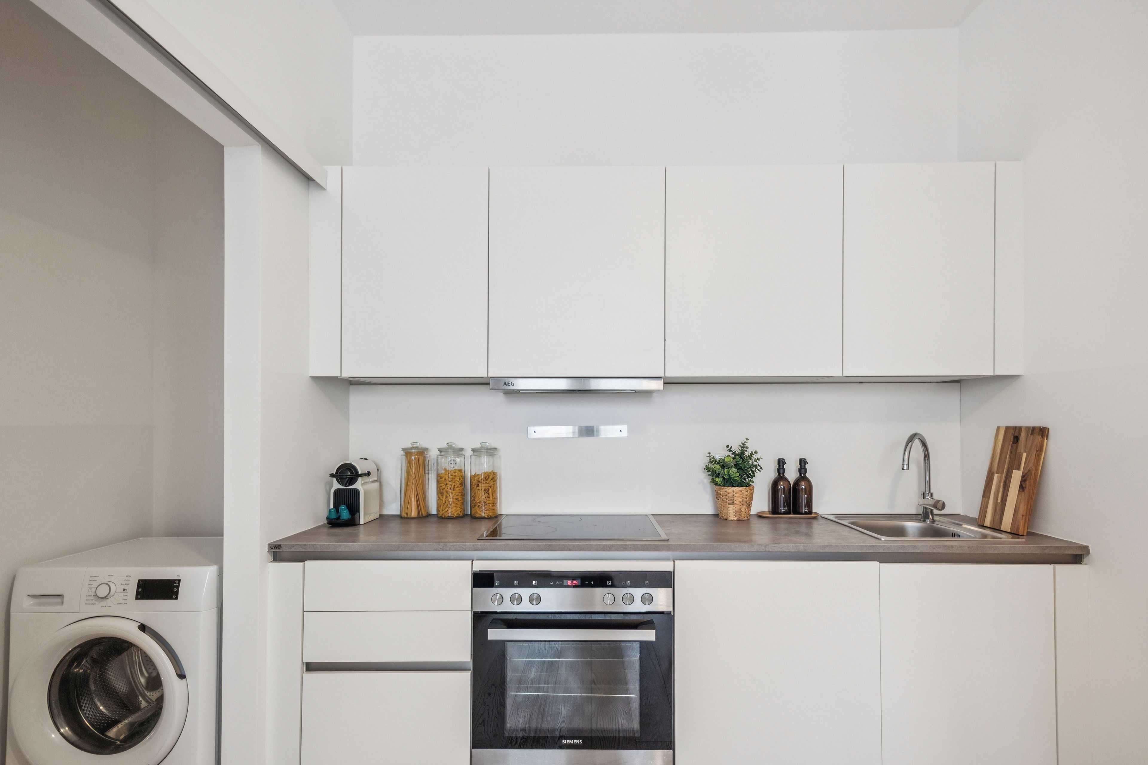 A compact kitchen with white cabinets, a built-in oven, a washing machine, and a stainless steel sink.