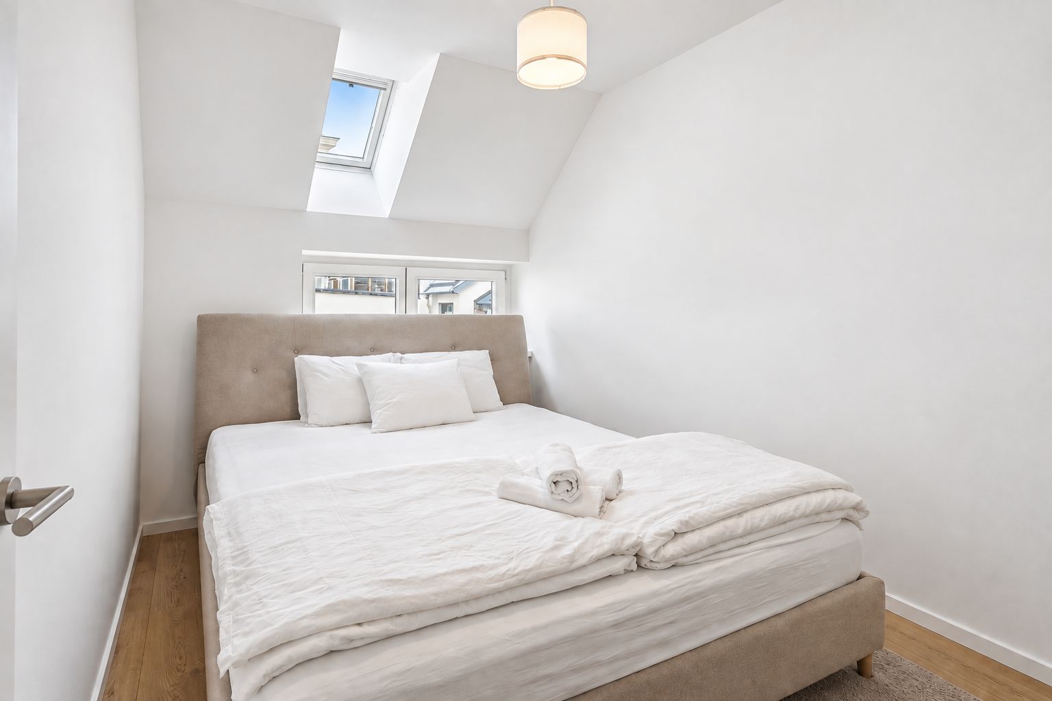 Een slaapkamer met een bed, kussens, een opgerolde handdoek, een raam en een dakraam.