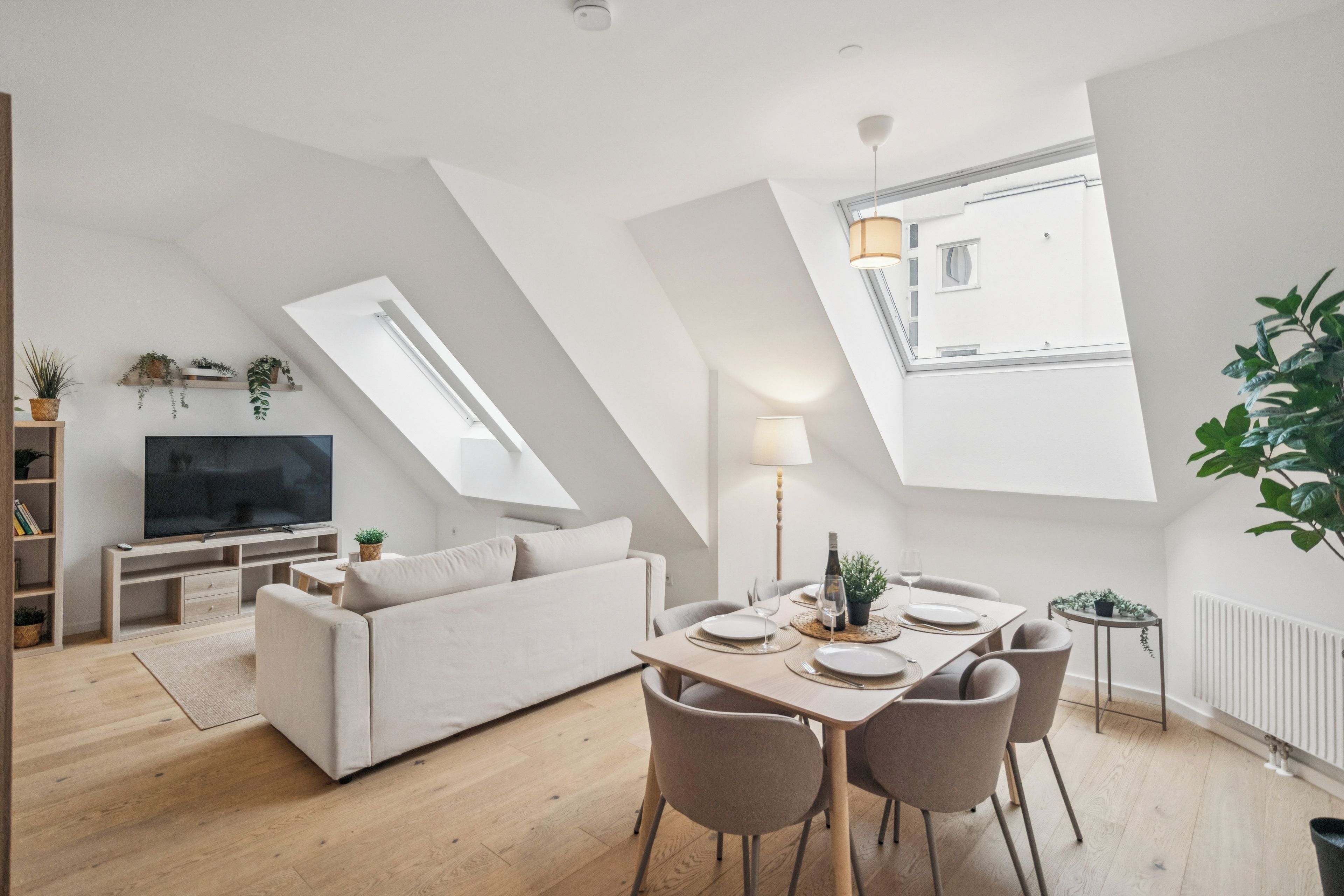 A modern loft-style living space with a dining area, a sofa, a TV, and a large window providing natural light.