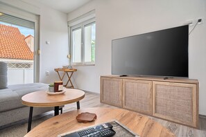 A living room with a TV on a wooden cabinet, a grey sofa, a round wooden table, and a window with a view of a red-tiled roof. - Spacious apartment with parking and big balcony, 4 min walk to beach (Split)