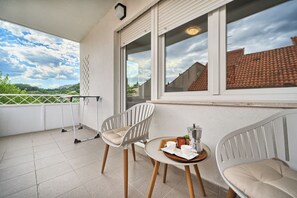 A balcony with two white chairs, a small round table with a tray of items, and a view of a red-tiled roof and greenery. - Spacious apartment with parking and big balcony, 4 min walk to beach (Split)