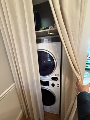 A compact, wall-mounted washing machine with a control panel and a circular door, partially obscured by beige curtains. - Luxury Furnished 2BR Condo, RH Design, Monthly Stays (Greensboro)