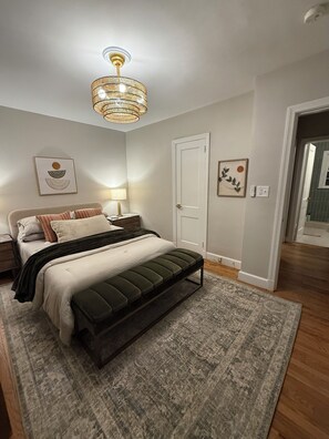 A bedroom with a bed, bedside table, lamp, and a hanging light fixture. - Luxury Furnished 2BR Condo, RH Design, Monthly Stays (Greensboro)