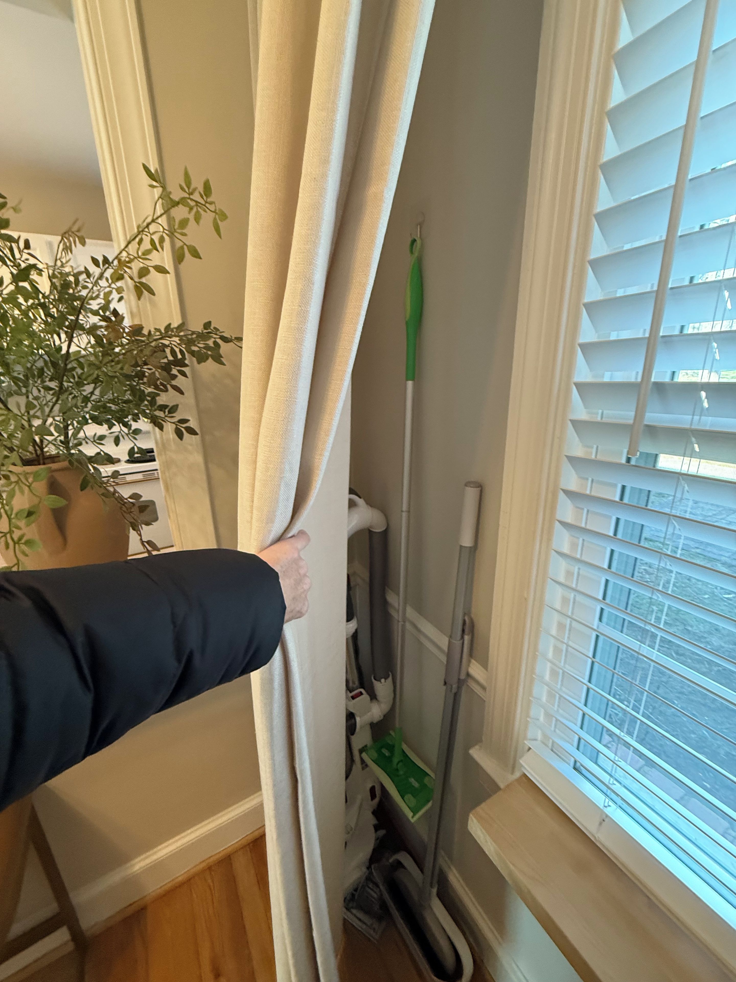 A hand holding a curtain, a mop, and a potted plant in a room with a window and blinds.