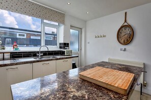 A kitchen with a granite countertop, a wooden cutting board, a vintage clock, and a window with a view of buildings. - Ammonite Cottage, Sea Views (Port Mulgrave)