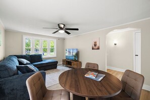 Ein Wohnzimmer mit einer Eckcouch, einem runden Holztisch mit Stühlen, einem Deckenventilator, einem Fernseher und einer Tür.