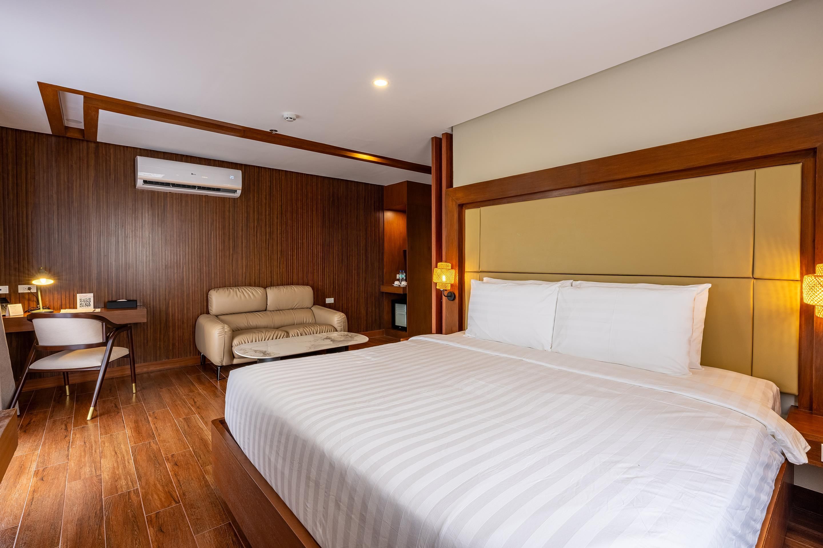 A hotel room with a large bed, a sofa, a desk with a chair, and a wall-mounted air conditioner.
