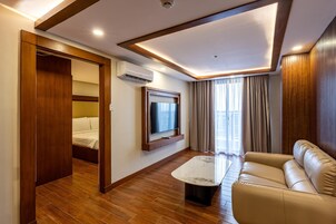 A hotel room with a wooden floor, a sofa, a TV mounted on the wall, and a bed with a wooden wardrobe.