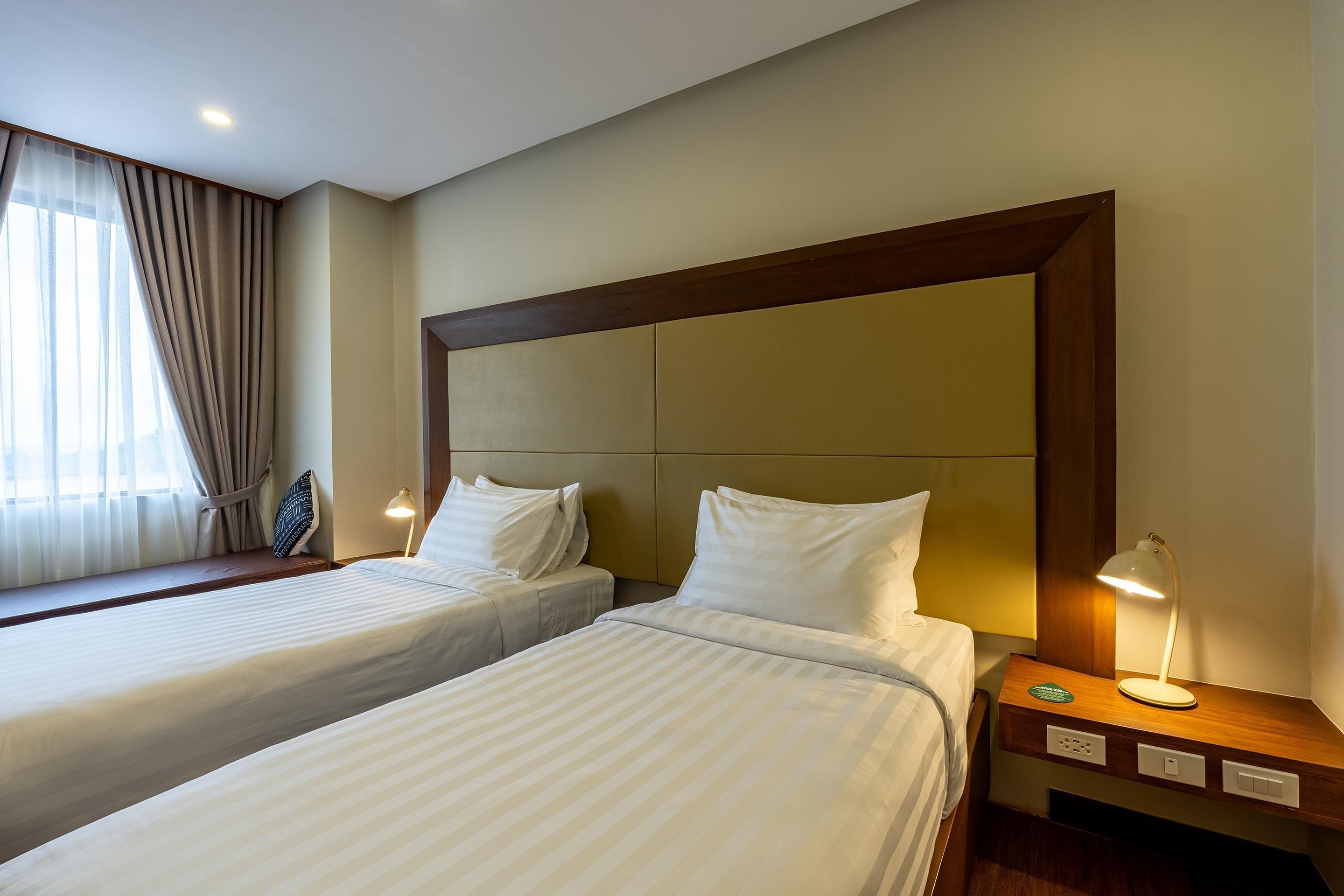 Habitación de hotel con dos camas, una ventana con cortinas, una mesita de noche con lámpara e iluminación en la pared.