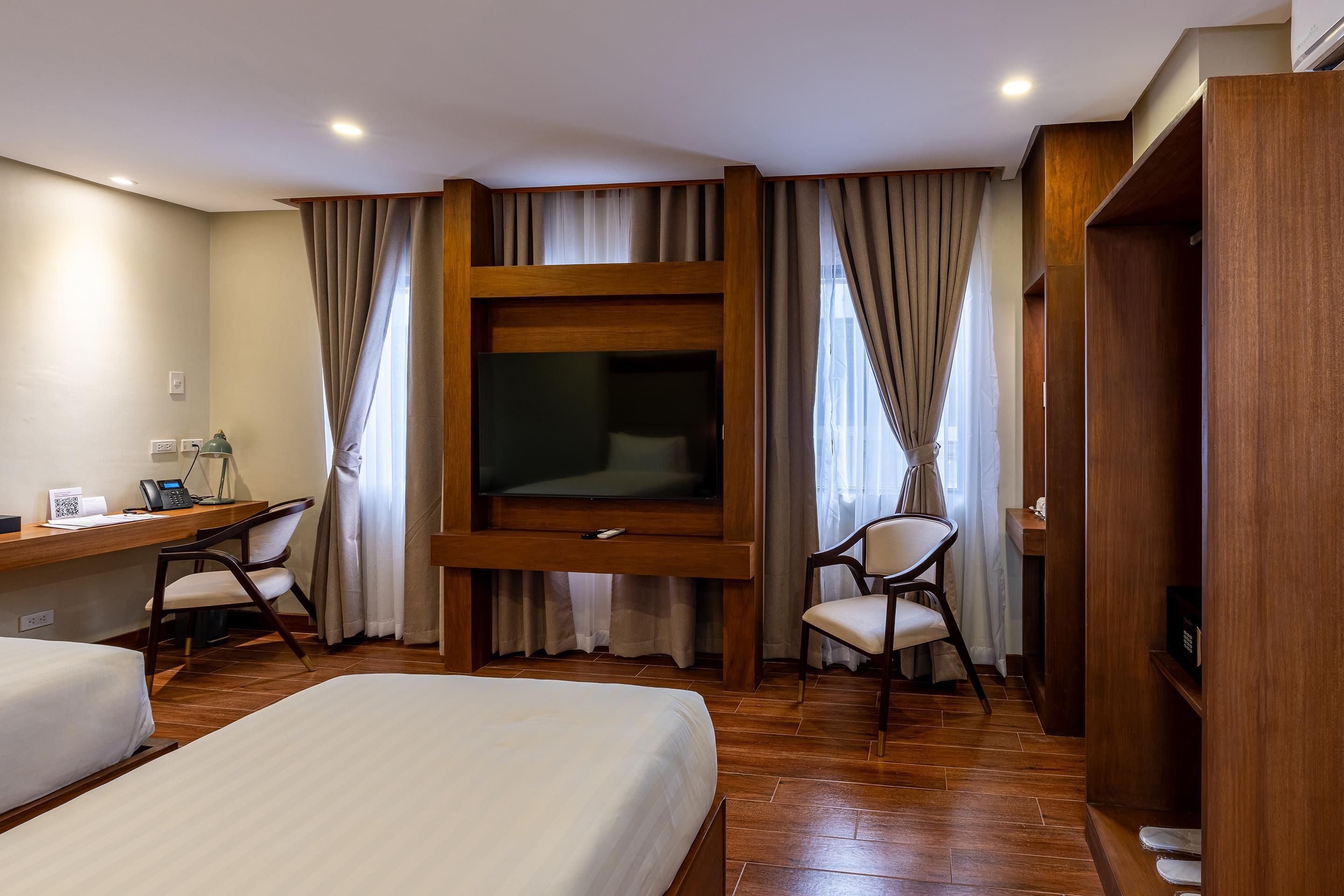 A hotel room with a bed, a desk with a telephone, a chair, and a television mounted on a wooden panel.