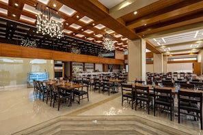 A spacious hotel dining area with multiple tables and chairs, a bar area with shelves of bottles, and a modern ceiling design with recessed lighting.