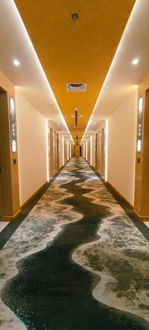 A hotel corridor with a central walkway, walls with vertical light fixtures, and a ceiling with recessed lighting.