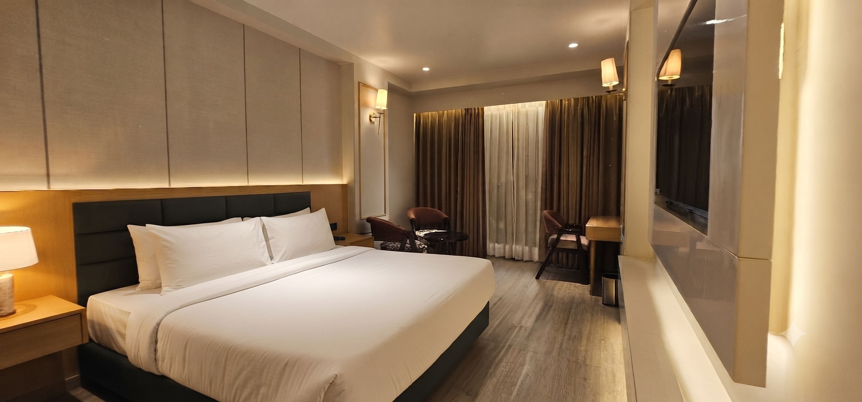 A hotel room with a large bed, a desk, two chairs, and a window with curtains.