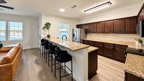 A modern kitchen with a central island, bar stools, and dark wooden cabinets. - Howdy House | Private Party Deck • 2.5 Miles to A&M (College Station)