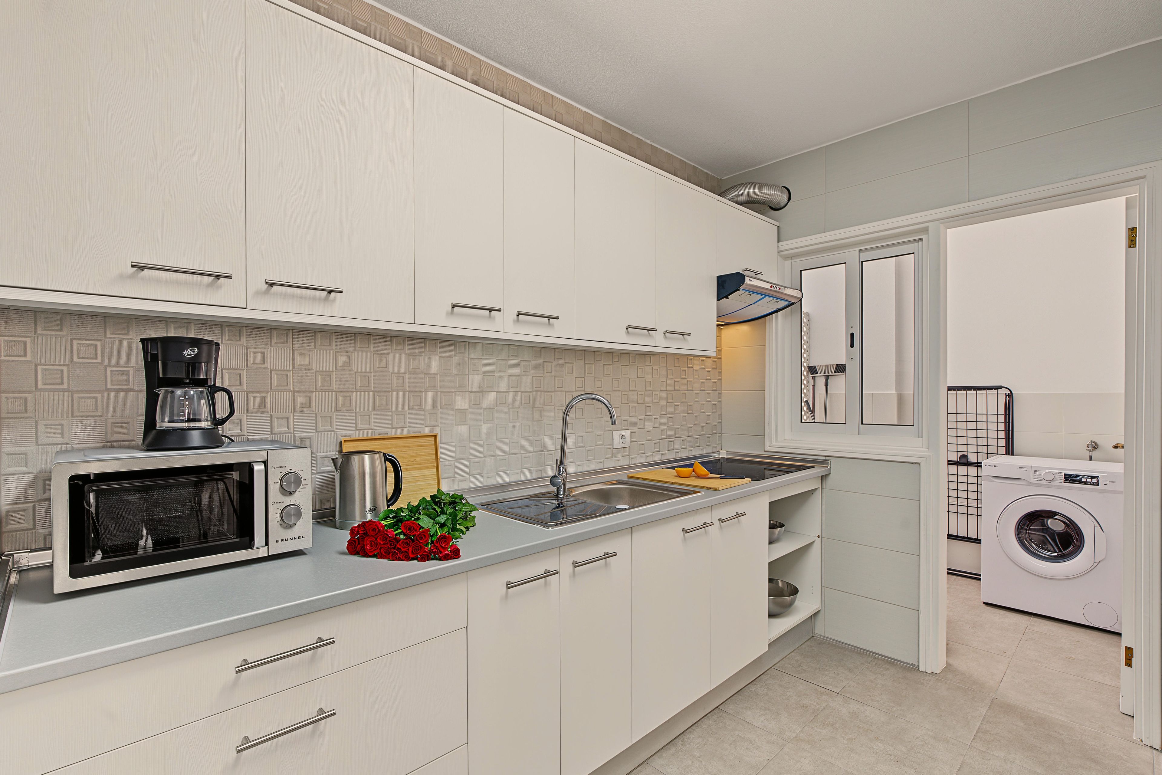 Une cuisine moderne avec des meubles blancs, un four à micro-ondes encastré, une machine à café et un lave-linge dans la pièce adjacente.