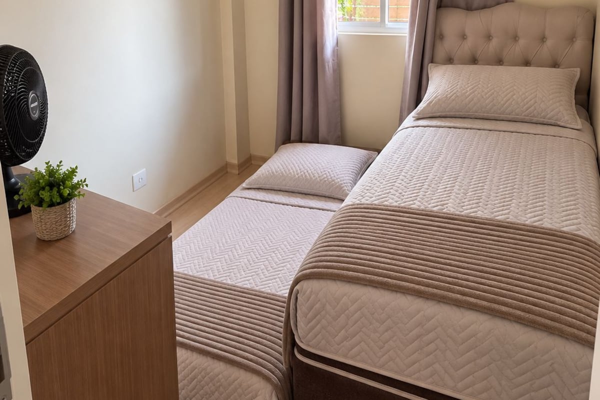 A bedroom with a bed, a bedside table with a fan, and a window with curtains.