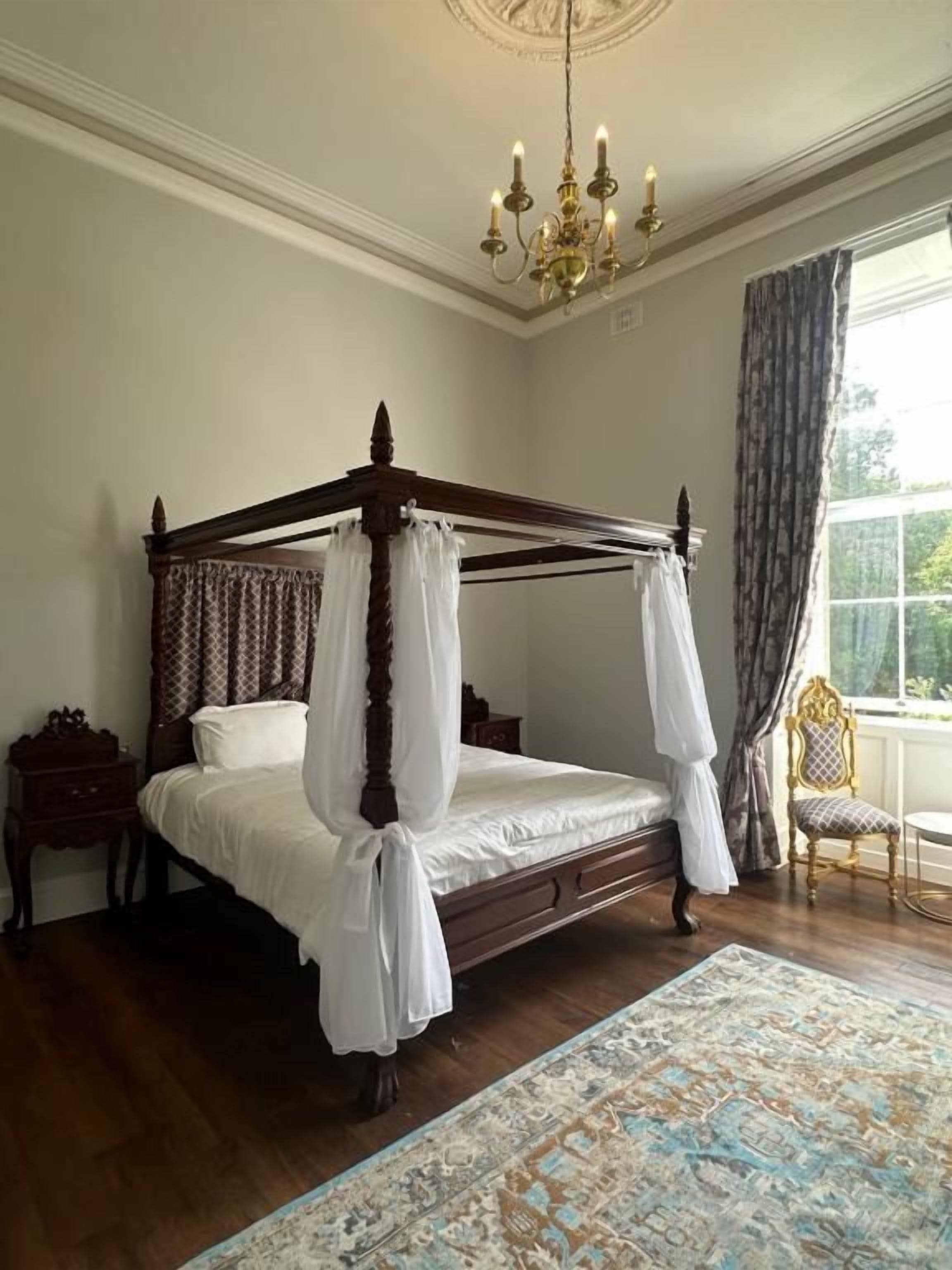 Ein Himmelbett mit Baldachin, eine Chandelüster, ein Fenster mit Vorhängen und ein gemustertes Teppich.