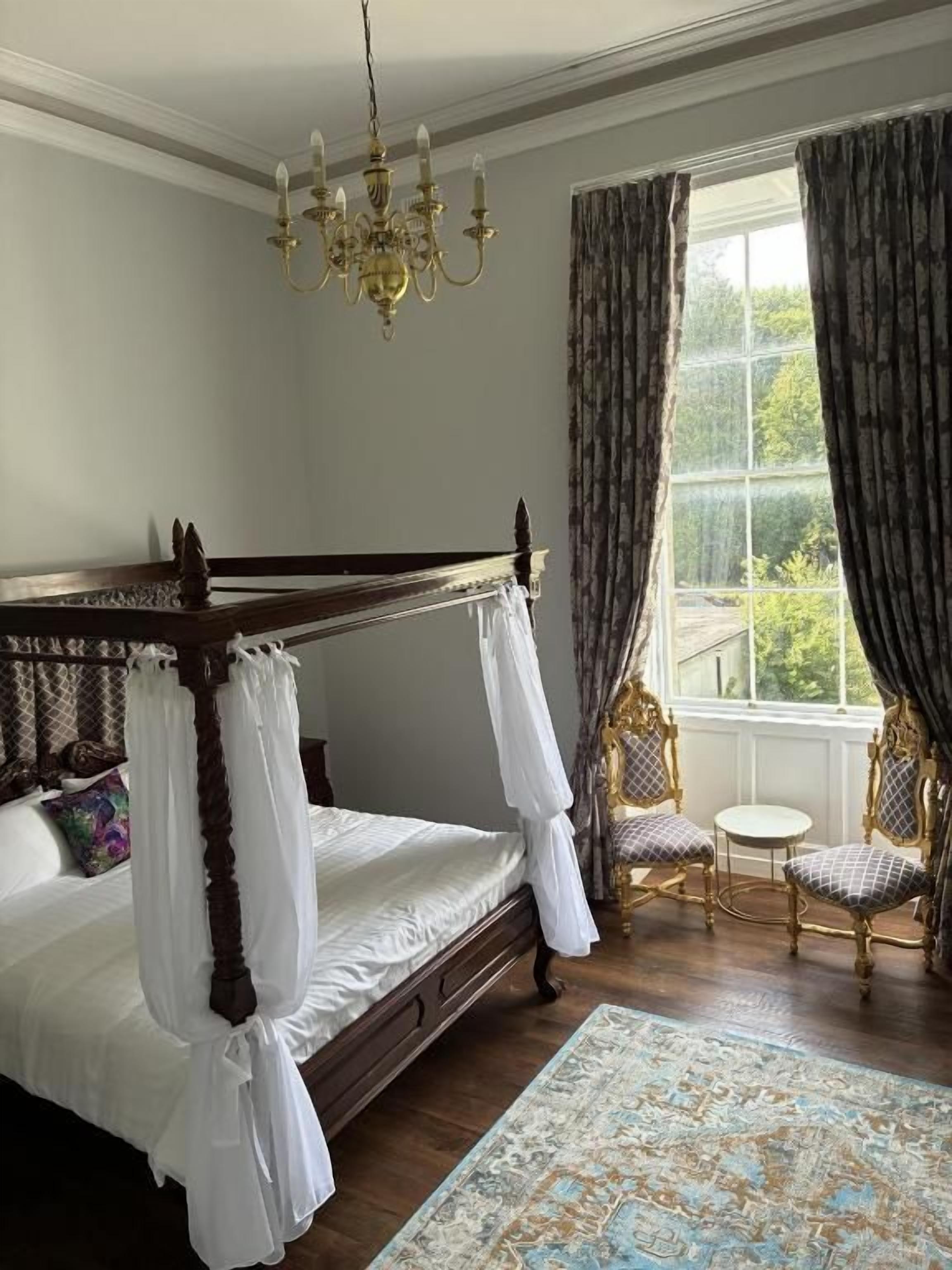 A four-poster bed with white linens, a chandelier, and a view of greenery through a window with curtains.