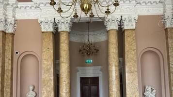 A grand hall with marble columns, a chandelier, and a door at the end.