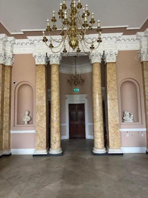A grand hall with marble columns, a chandelier, and a door at the end.