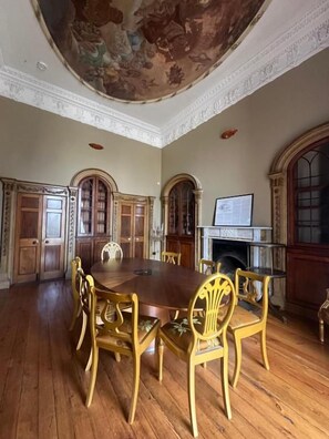 Uma sala de jantar com uma grande mesa de madeira e cadeiras, lareira e um afresco no teto.