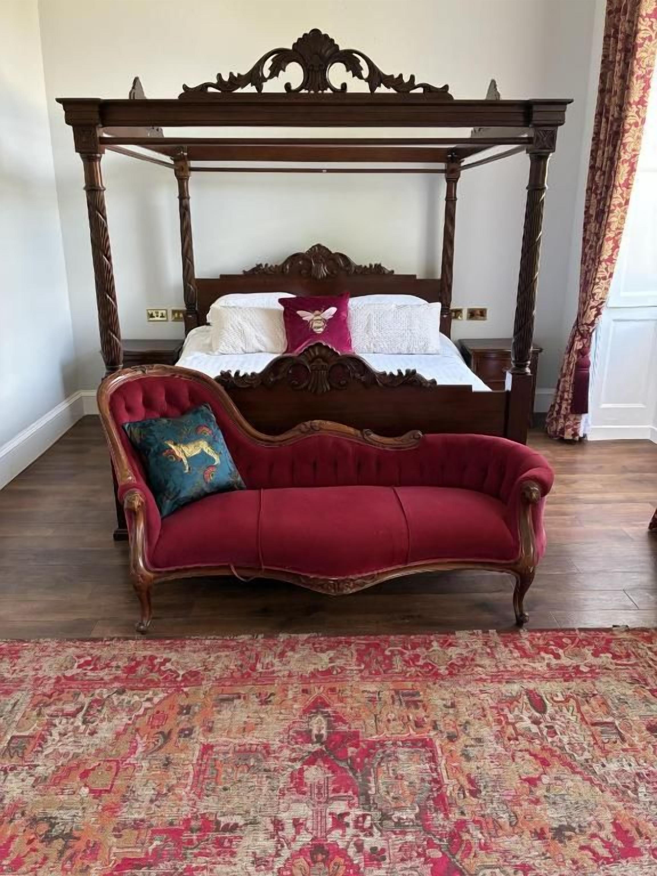 A four-poster bed with a red upholstered sofa in front, a patterned rug, and a wooden floor.