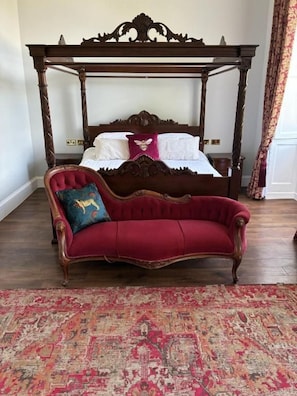 A four-poster bed with a red upholstered sofa in front, a patterned rug, and a wooden floor.