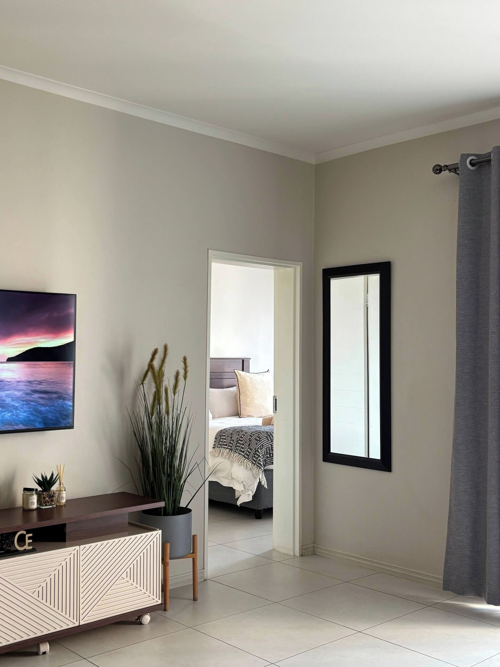 A modern living room with a large TV, a console table, a potted plant, and a view into a bedroom.