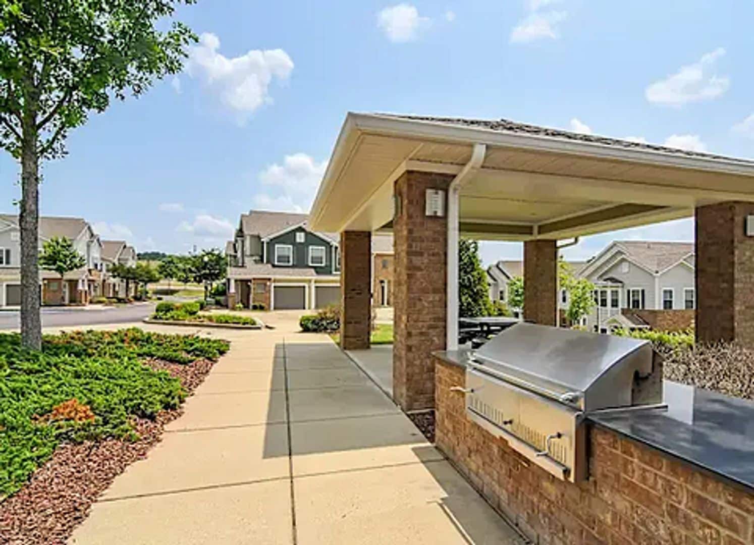 A residential area with a barbecue grill under a covered patio, a sidewalk, and houses in the background.