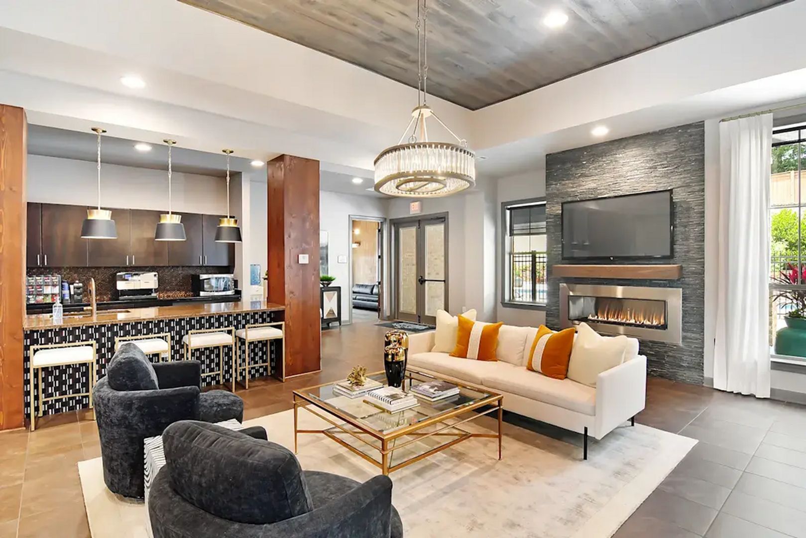 A modern living room with a sofa, coffee table, and chairs. There is a kitchen area with a bar and pendant lights. A fireplace is visible, and a TV is mounted on the wall.