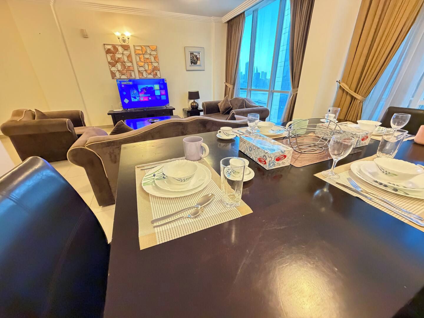 Un comedor con una mesa preparada para cuatro personas, un televisor al fondo y vistas de la ciudad a través del ventanal.