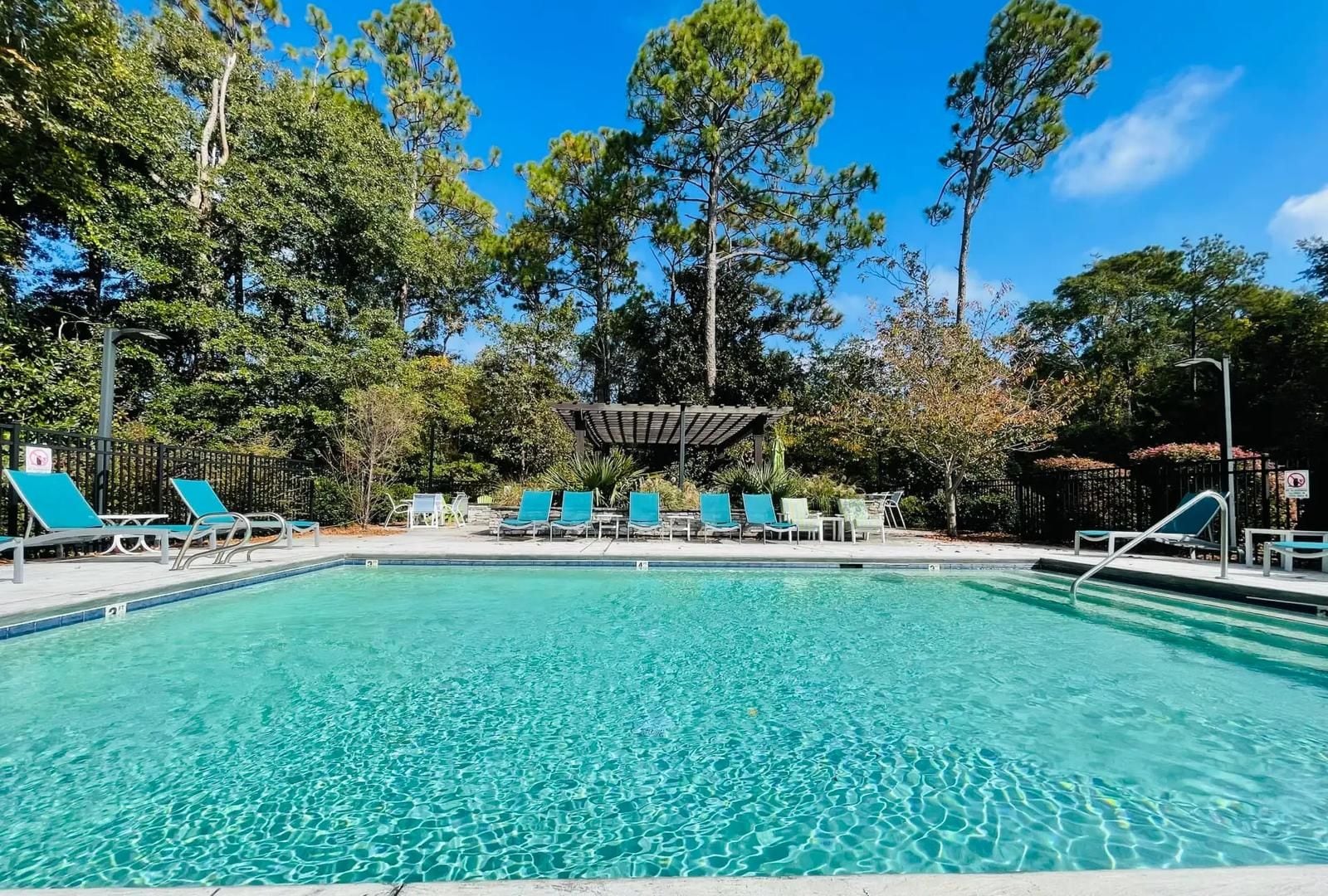 A clear swimming pool with lounge chairs and a shaded area, surrounded by trees.
