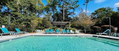 A clear swimming pool with lounge chairs and a shaded area, surrounded by trees.
