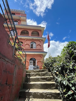 A multi-story building with a staircase leading to the entrance, surrounded by greenery. - Innate Pension Guest House and café  (Dhulikhel)