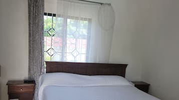 A bedroom with a wooden bed, a nightstand, a window with a curtain, and a view of greenery outside.