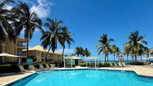 Una piscina azul y cristalina rodeada de palmeras y sillas de descanso, con un edificio al fondo.