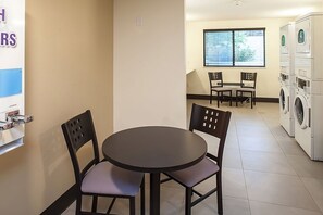 Ein Zimmer mit einem runden Tisch, vier Stühlen, einem Fenster mit Jalousien und einer Waschmaschine im Hintergrund.