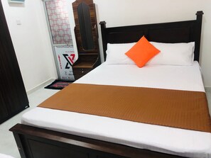Ein ordentlich bezogenes Bett mit brauner Tagesdecke und einem orangefarbenen Kissen, ein Holz-Kopfteil und ein kleiner Nachttisch mit einer Lampe.