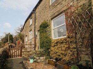 A stone house with a wooden gate, a small garden with plants, and a clear sky. - Crag Cottage (Settle)