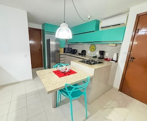 A compact kitchen with teal cabinets, a gas stove, a refrigerator, and a dining area with turquoise chairs. - Marupiara Muro Alto (Ipojuca)
